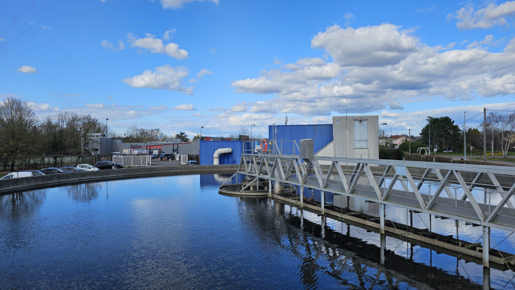Station d'épuration avec bassin de décantation sous un ciel bleu, entourée de bâtiments et d'arbres.