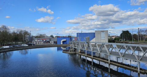 Station d'épuration avec bassin de décantation sous un ciel bleu, entourée de bâtiments et d'arbres.