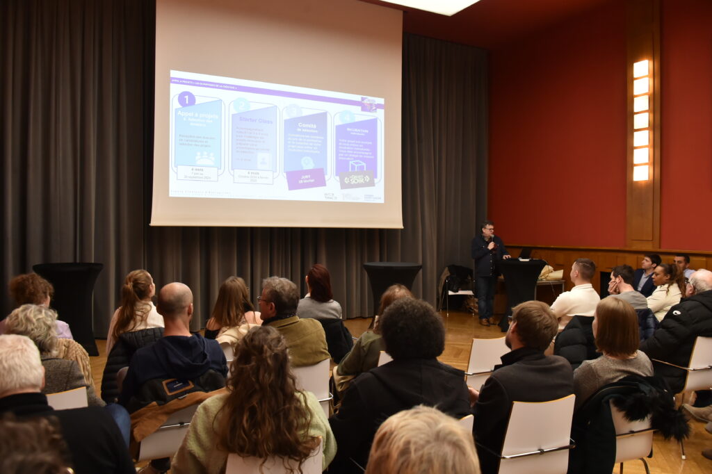 Conférence en salle avec présentation PowerPoint sur un appel à projets, public attentif et orateur au micro.