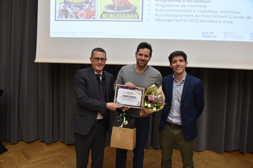 Trois hommes posent avec un certificat lors d'une remise de prix, l'un tenant un bouquet de fleurs.