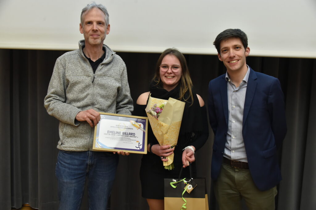 Trois personnes posent avec un prix, une femme au centre tenant des fleurs et un homme avec un diplôme à gauche.