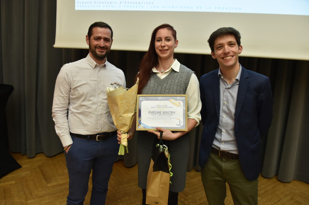 Trois personnes posent pour une photo lors d'une cérémonie de remise de prix, souriantes avec un certificat et des fleurs.