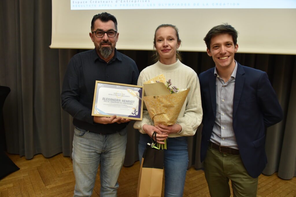 Groupe de trois personnes lors d'une remise de prix, tenant un diplôme et un bouquet de fleurs.