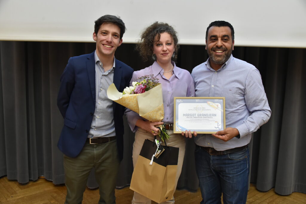 Trois personnes souriantes lors d'une remise de prix, avec un bouquet de fleurs et un certificat de récompense.
