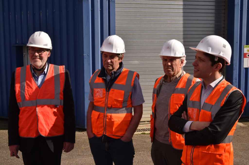 Quatre personnes en uniforme de sécurité avec casques et gilets orange devant un bâtiment industriel bleu.