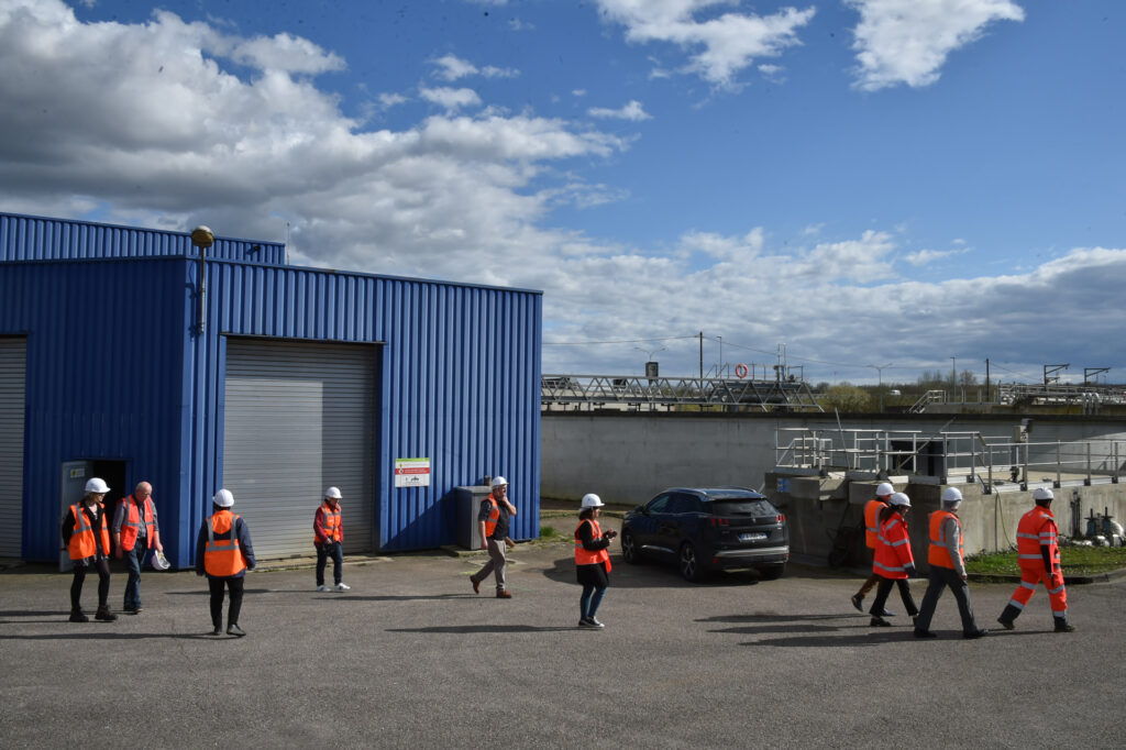 Personnes en gilets orange et casques blancs visitant un site industriel près d'un bâtiment bleu sous un ciel nuageux.