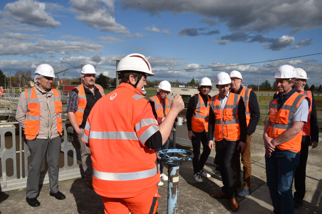 Groupe de travailleurs avec gilets orange et casques blancs lors d'une visite de chantier en plein air par temps ensoleillé.