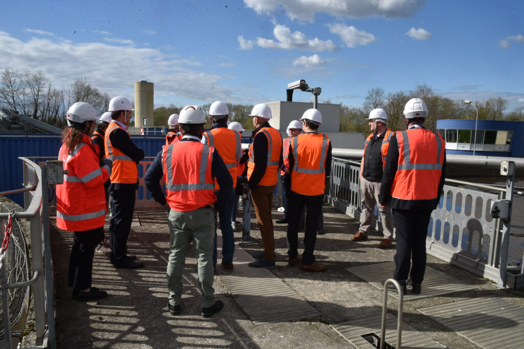 Personnes en visite de chantier avec gilets orange et casques, discutant à l'extérieur sous un ciel ensoleillé.