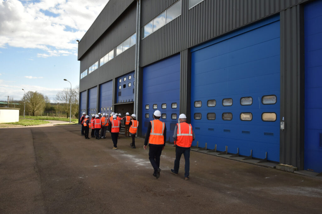 Personnes en gilets orange et casques blancs entrant dans un bâtiment industriel bleu sous un ciel dégagé.