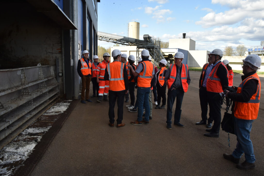 Groupe en visite industrielle avec casques et gilets orange, devant une usine sous un ciel en partie nuageux.