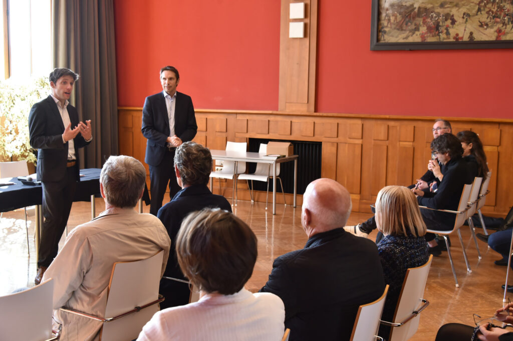 Conférence en salle avec deux orateurs en costume s'adressant à un public attentif, dans un environnement professionnel.