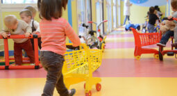 Enfants jouant avec des chariots en plastique et des tricycles colorés dans une crèche, sous la supervision d'un adulte.
