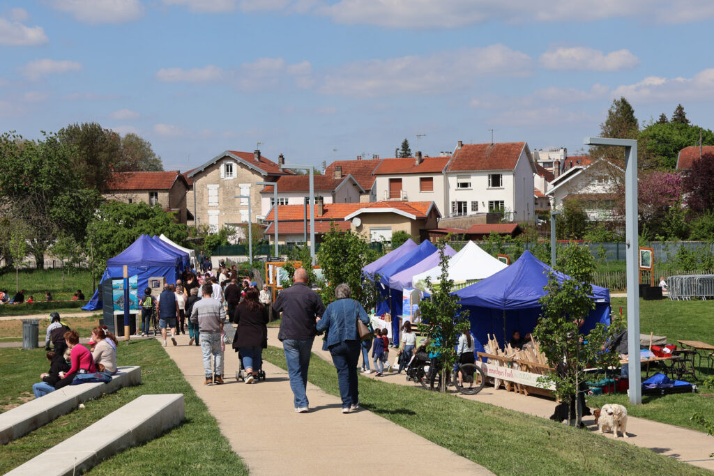 Passants dans un parc lors d'un marché en plein air avec des stands de tentes bleues sous un ciel ensoleillé.