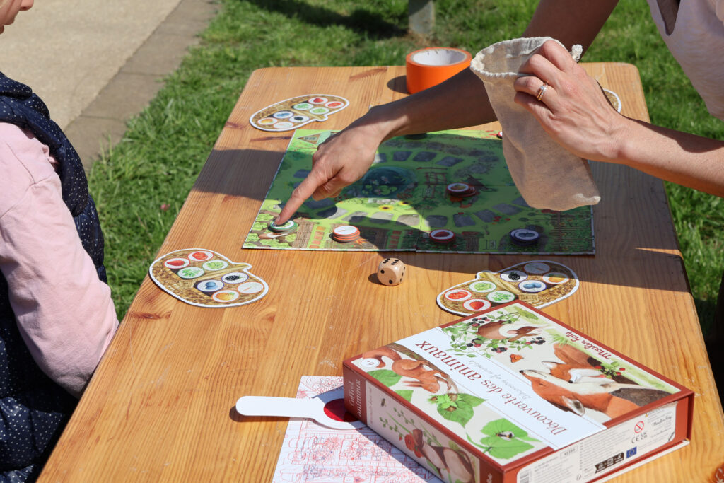 Personnes jouant à un jeu de société éducatif en plein air avec des cartes et des pions sur une table en bois.