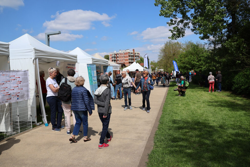 Événement en plein air avec des stands informatifs et des visiteurs sous un ciel bleu, dans un cadre verdoyant.