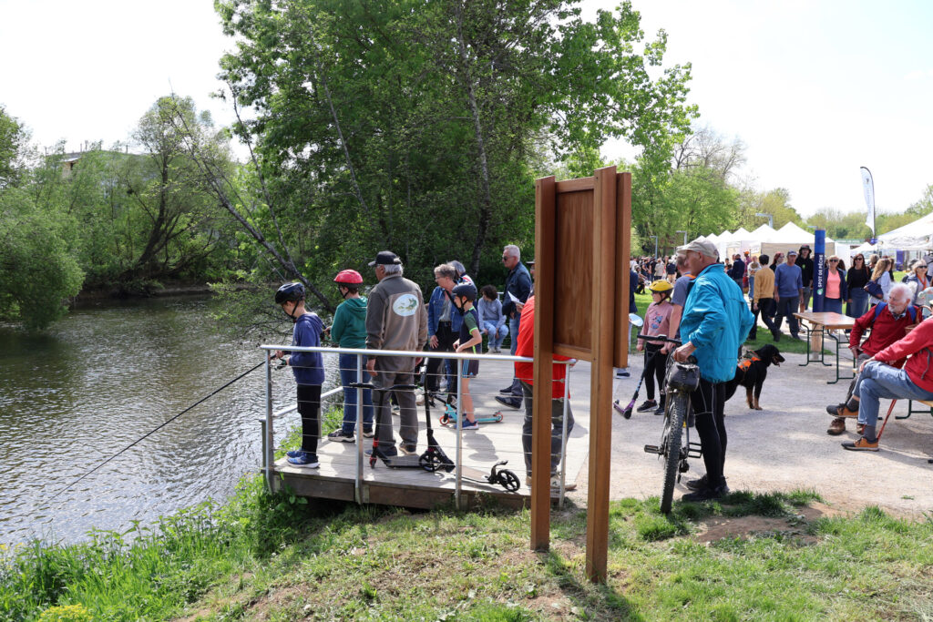 Des personnes assistent à un événement en plein air près d'une rivière, entourées d'arbres et de tentes, sous un ciel dégagé.