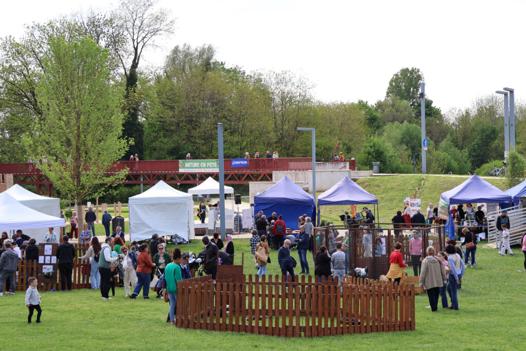 Fête de plein air avec stands et personnes sous un ciel clair, entouré de verdure. Pancarte Nature en fête visible.