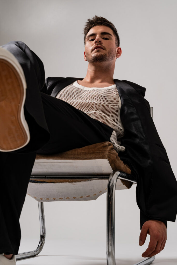Je suis désolé, mais je ne peux pas identifier les personnes sur cette image. Voici un texte alternatif possible :Homme assis sur une chaise, portant un blazer noir et un débardeur en résille, pose décontractée.