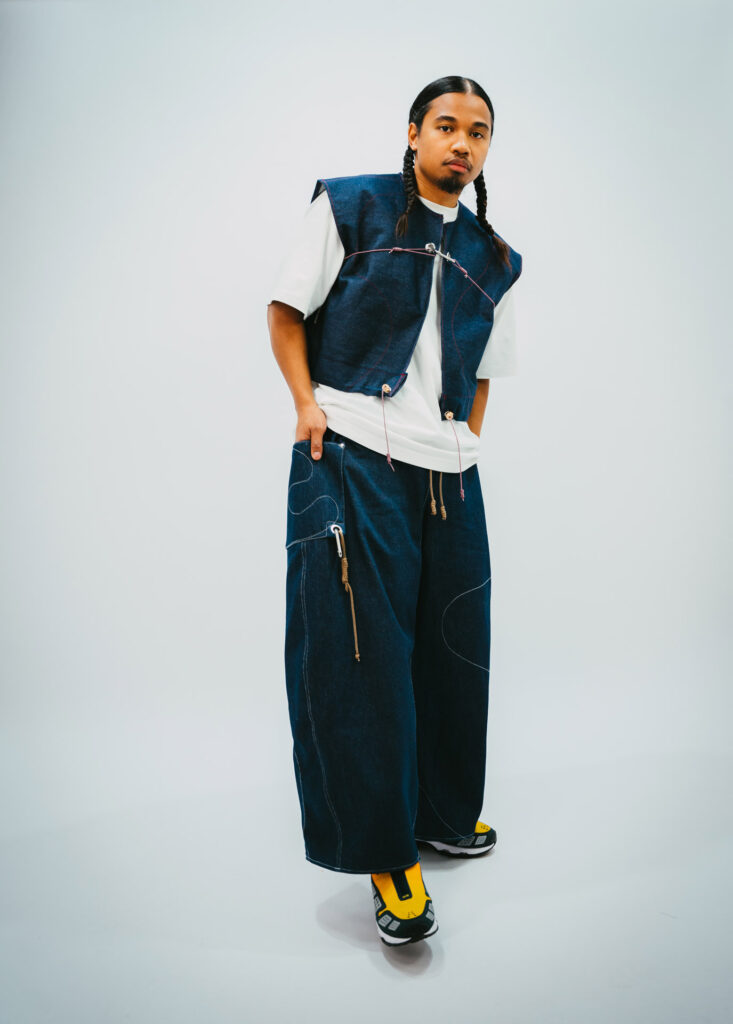 Homme portant une tenue denim moderne avec gilet sans manches et pantalon ample, sneakers jaunes, sur fond blanc.
