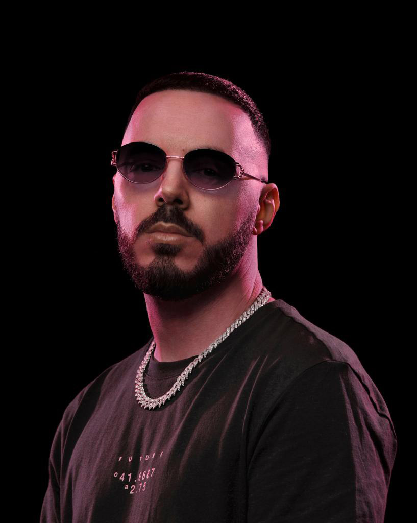 Bien sûr! Voici un texte alternatif :Homme à lunettes de soleil, barbe et chaîne argentée sur fond noir.