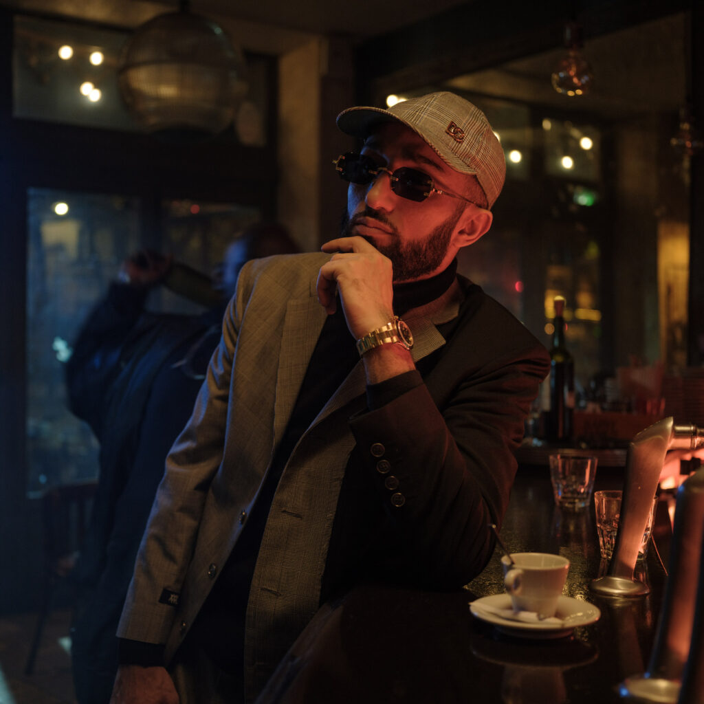 Un homme élégant avec des lunettes et une casquette réfléchit dans un café. Ambiance tamisée et chaleureuse.