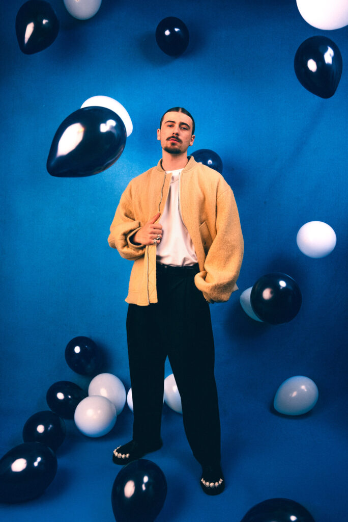 Homme en manteau beige debout parmi des ballons noirs et blancs sur fond bleu. Ambiance moderne et artistique.